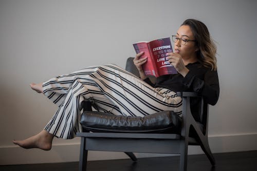 Top Feminino De Mangas Compridas Pretas, Calças Listradas Brancas E Pretas Lendo Livro Sobre Poltrona Acolchoada Com Moldura De Madeira Cinza