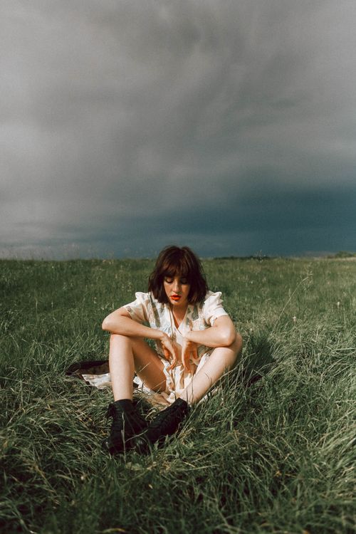 Free Woman sitting in a grassy field under a cloudy sky, wearing black boots. Stock Photo