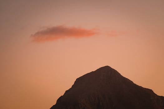 A stunning mountain silhouette during sunset with a warm orange sky and a lone cloud, capturing serene beauty.