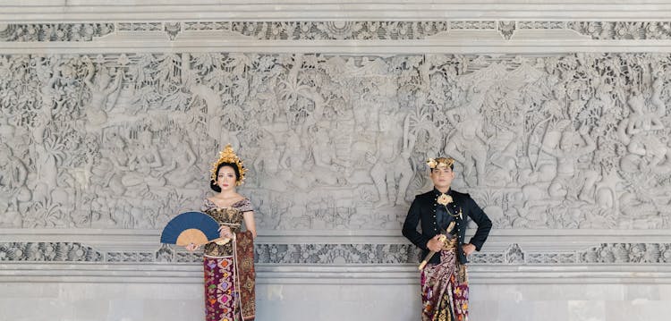 Man And Woman Wearing Traditional Clothes Standing Beside A Wall With Carving