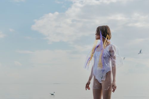 Free Stylish woman in sheer outfit standing on a serene beach under a clear sky. Stock Photo