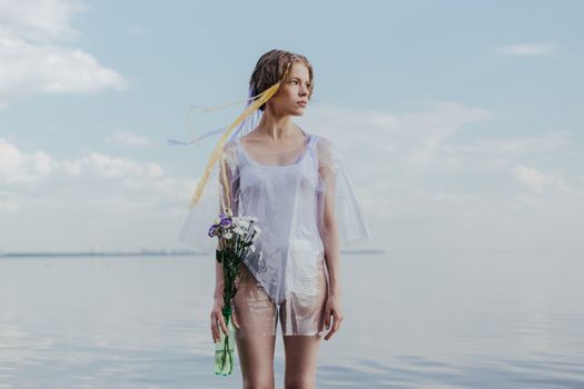 A woman in a sheer outfit stands by the ocean holding flowers, under a bright blue sky.