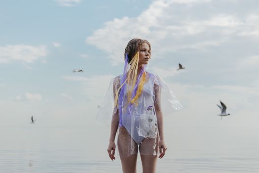 Stylish woman in a clear outfit standing by the sea under a blue sky.