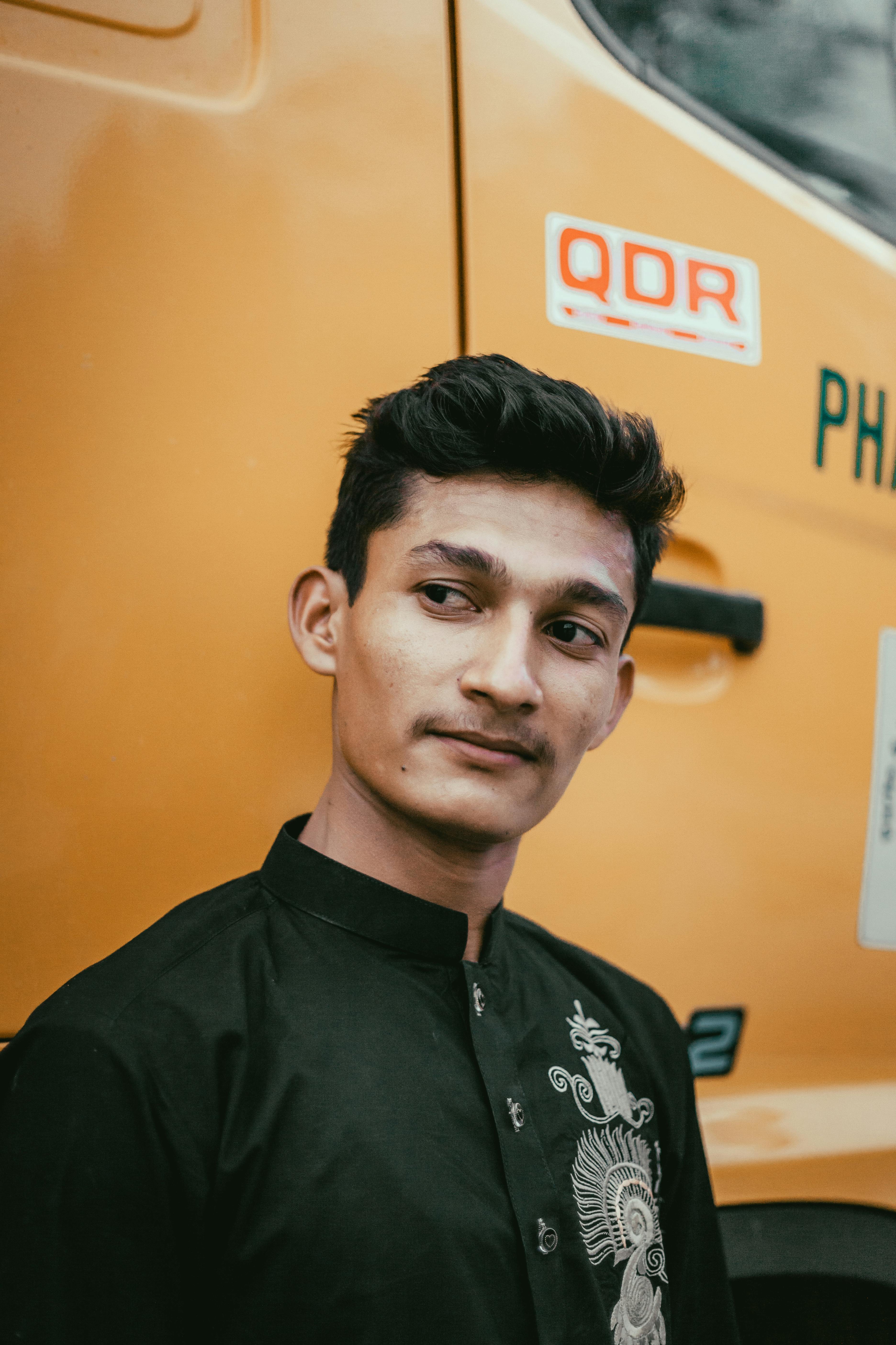 A young man poses confidently against a yellow truck, wearing a stylish black shirt.