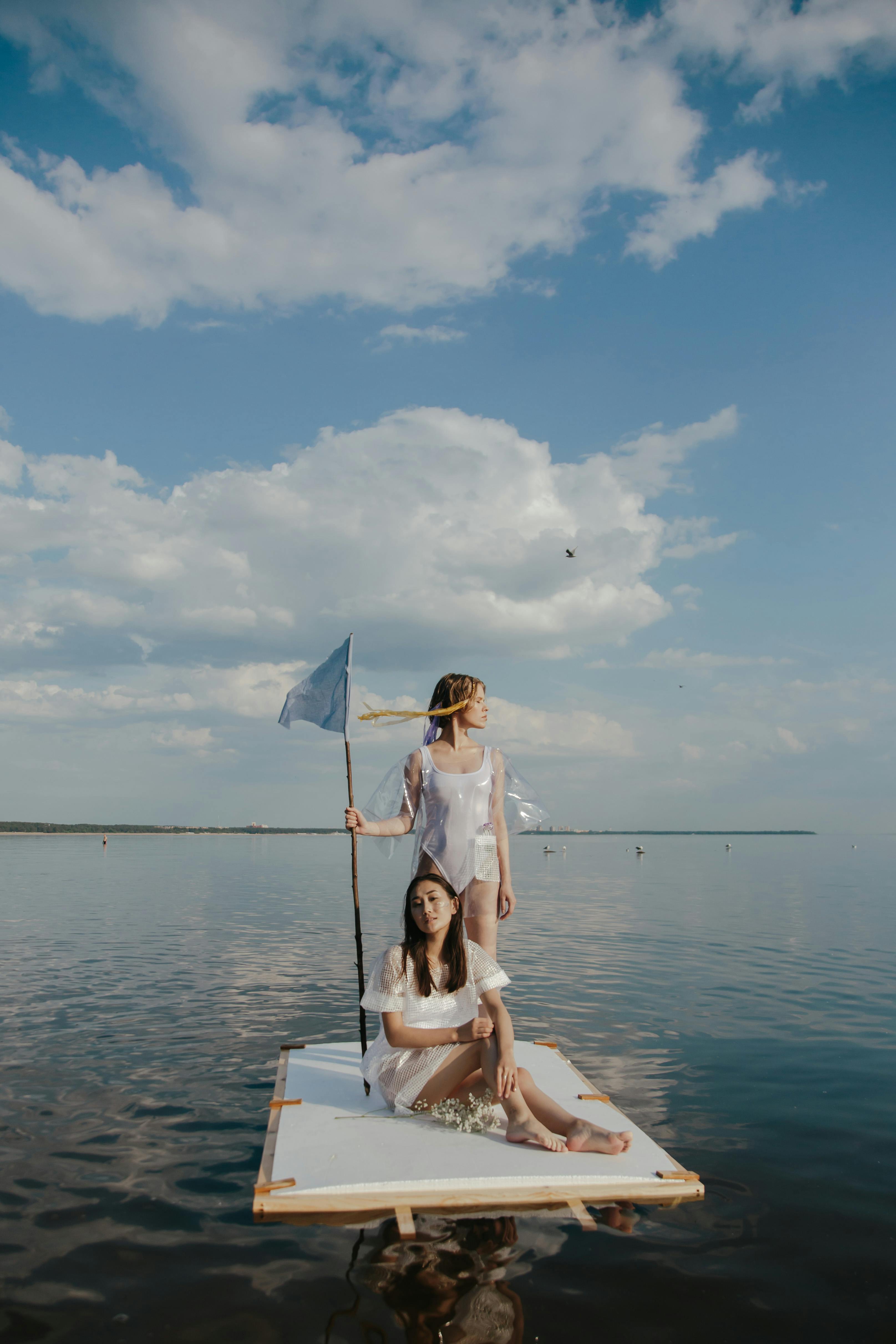 Two women on a raft in tranquil waters holding a flag under a clear sky.