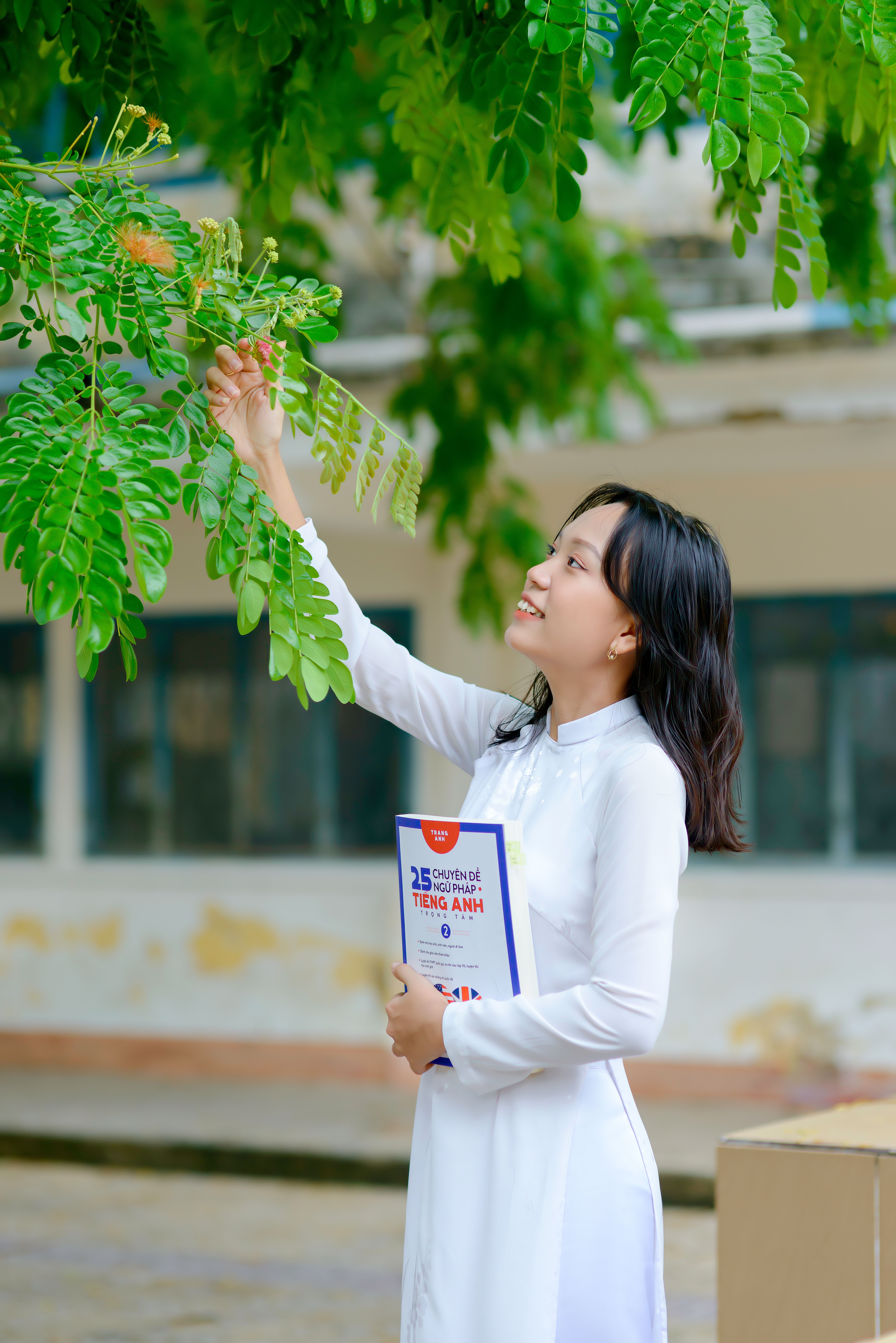 A young woman in a white dress holding a book reaches for leaves outdoors during the day.