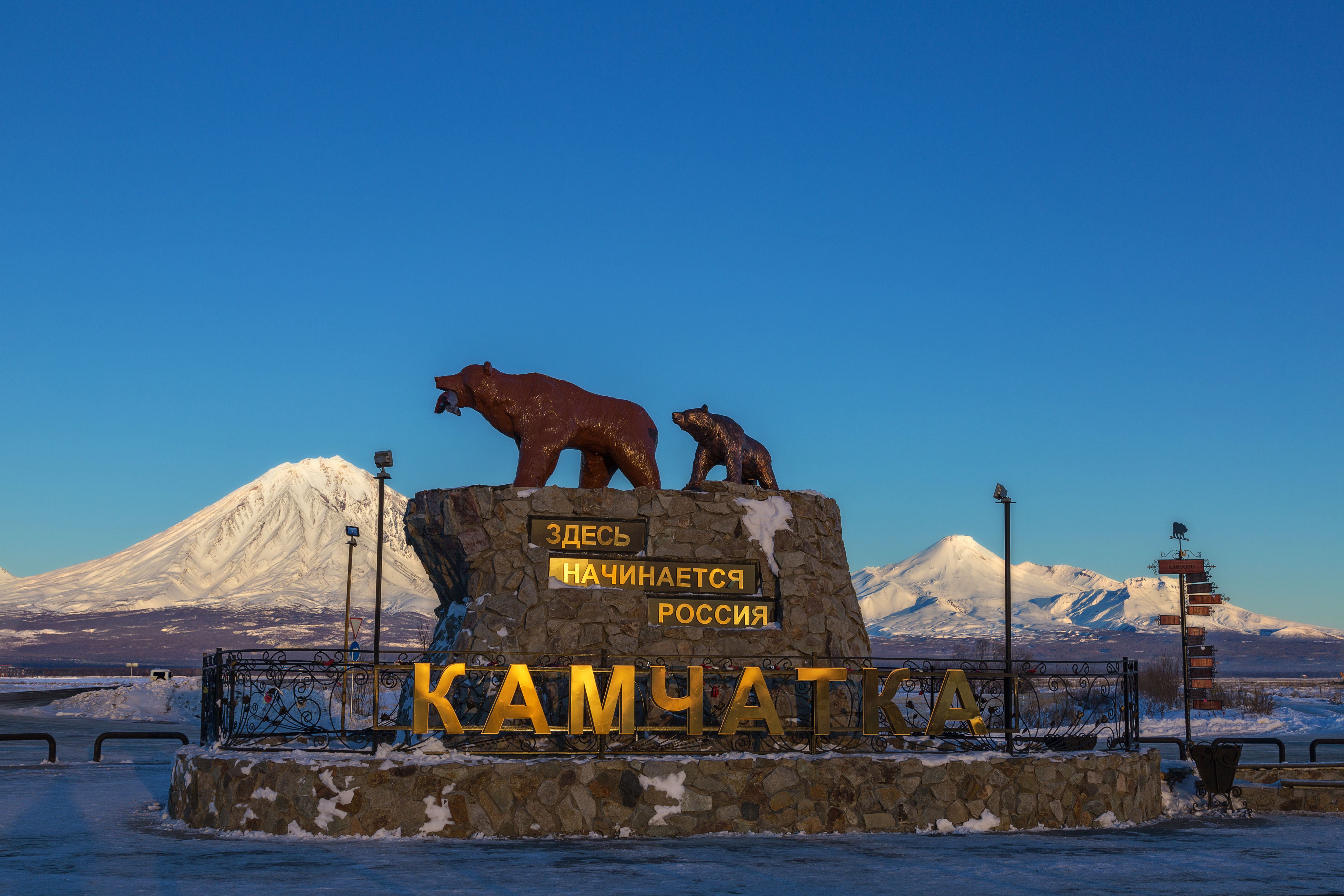 Kamchatka monument with bear sculptures against snow-covered mountains in clear blue sky.