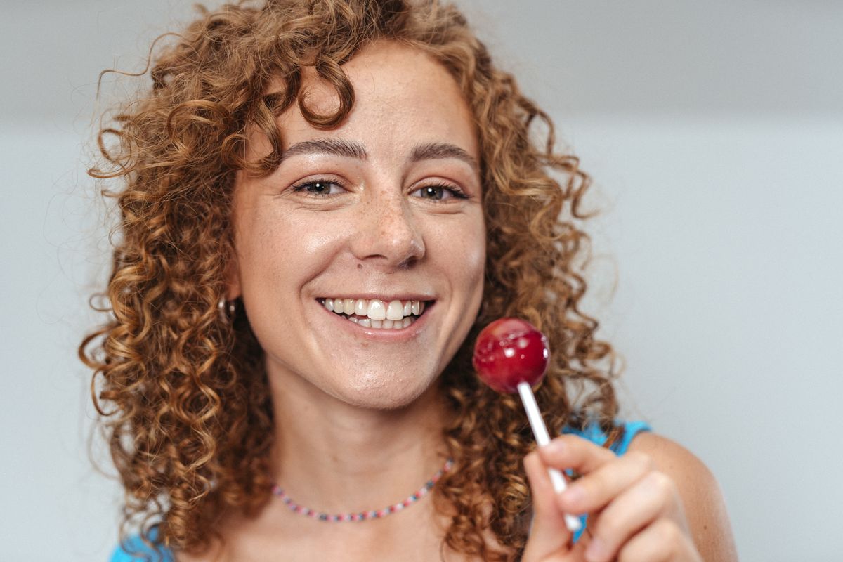 Happy woman with curly hair holding a red lollipop, smiling warmly.
