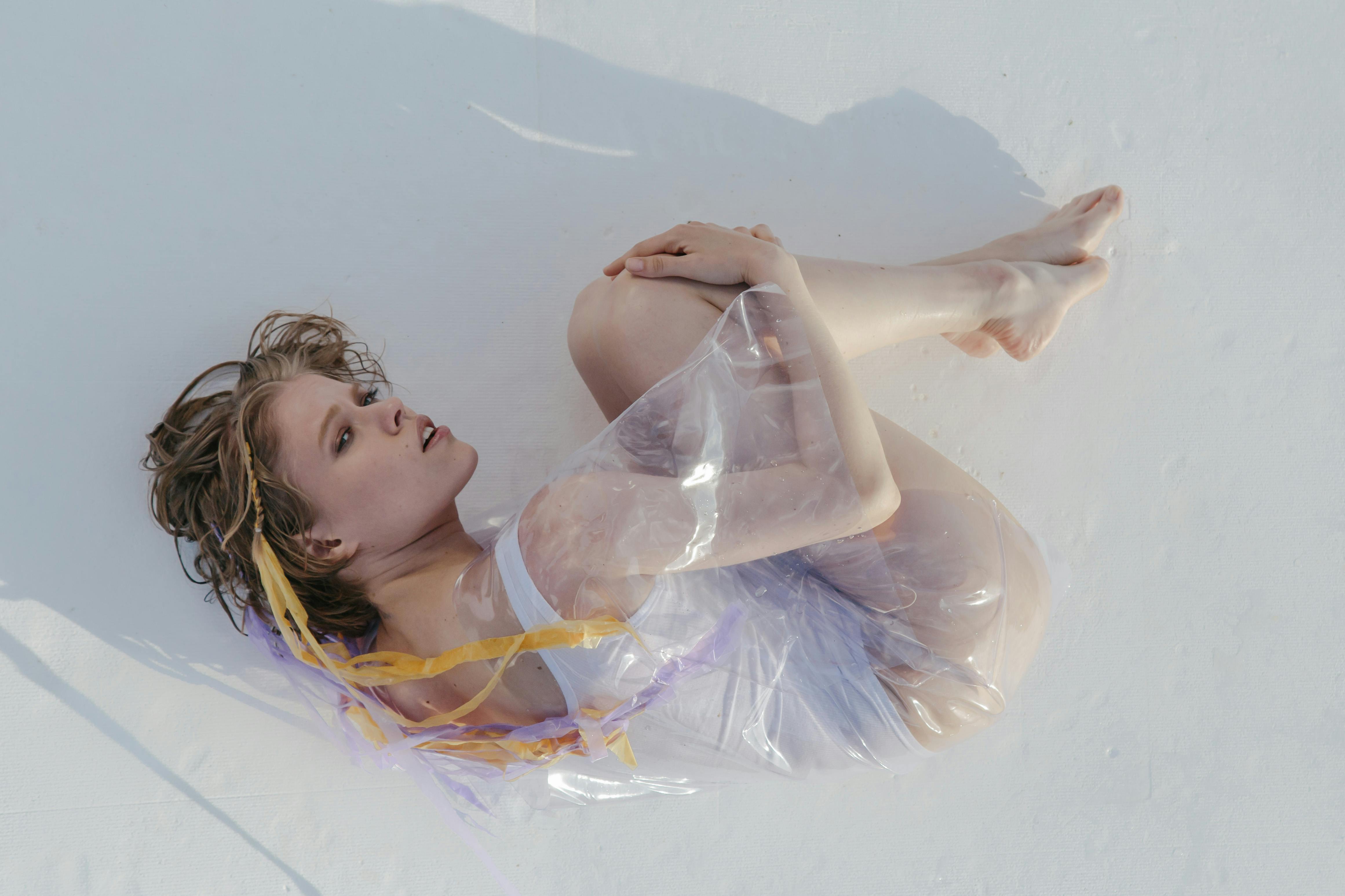 Creative image of a woman wrapped in plastic on a white surface, captured from above.