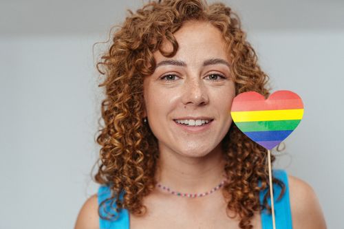 Free A cheerful woman with curly hair holds a rainbow heart sign, symbolizing pride and inclusivity. Stock Photo