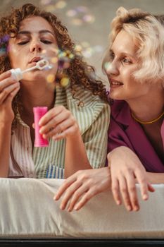 Two women friends enjoy blowing bubbles indoors, capturing a moment of happiness and togetherness.
