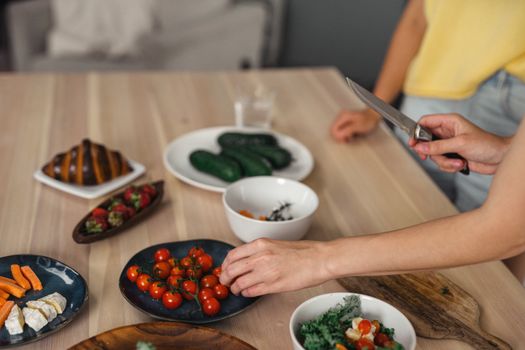 Hands slicing and arranging fresh vegetables and fruits on wooden table indoors.