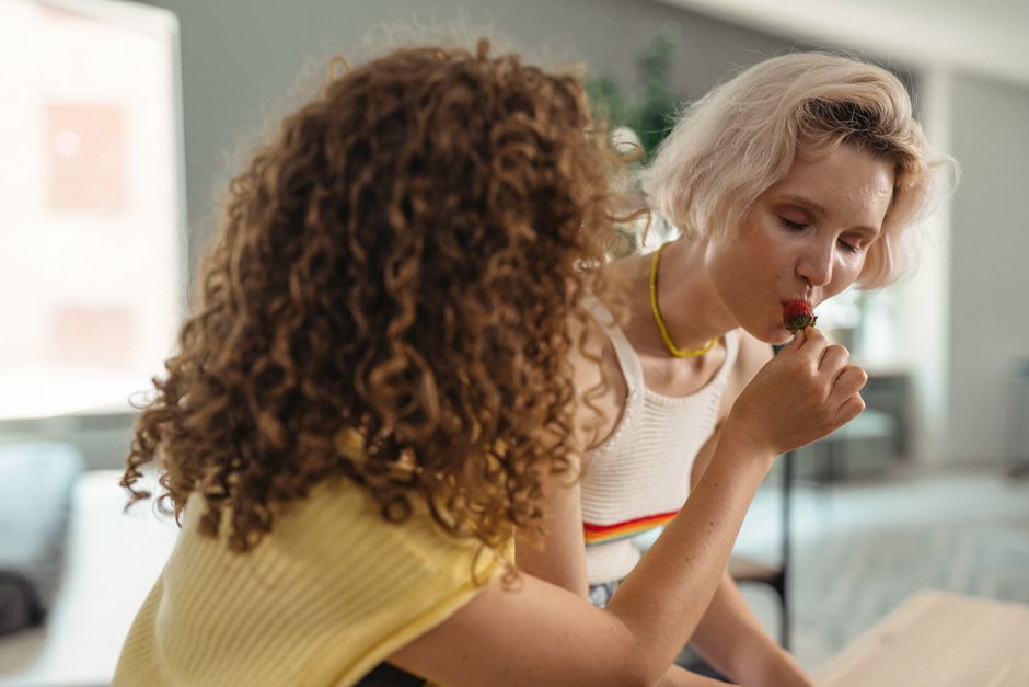 Two women enjoy a playful moment indoors, sharing strawberries and laughter, symbolizing friendship and closeness.