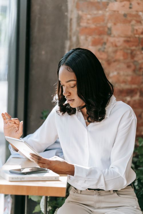 Free A woman in a white shirt sits by a window with a tablet, focused on her work. Stock Photo