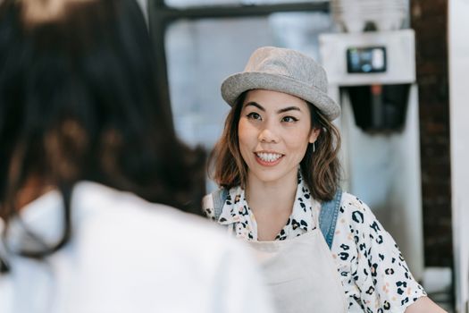 A friendly woman wearing a hat and apron smiles while interacting at a cafe.