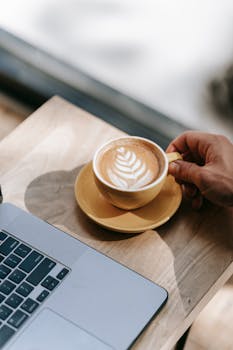 A hand holding a latte in a yellow cup with latte art beside a laptop on a wooden table.