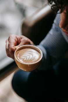 A person holding a cappuccino with intricate leaf latte art in a cozy setting.