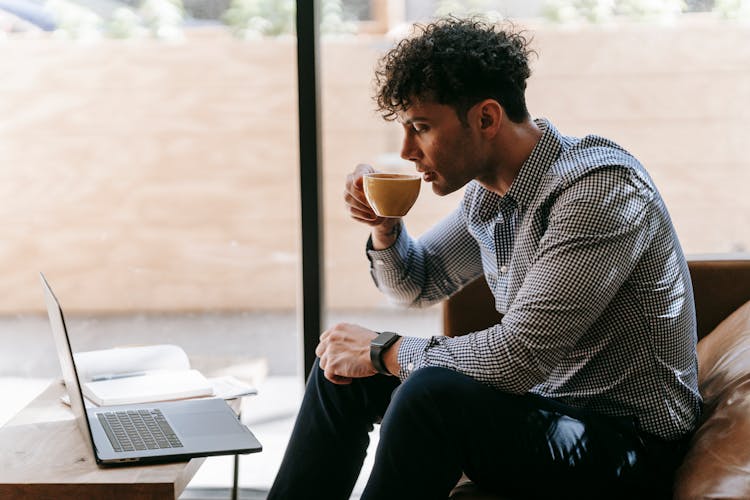 A Man Sipping Coffee While Sitting In Front Of A Laptop