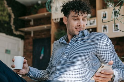 Free Man with curly hair sits indoors, stylishly looking at his phone while holding a coffee. Stock Photo