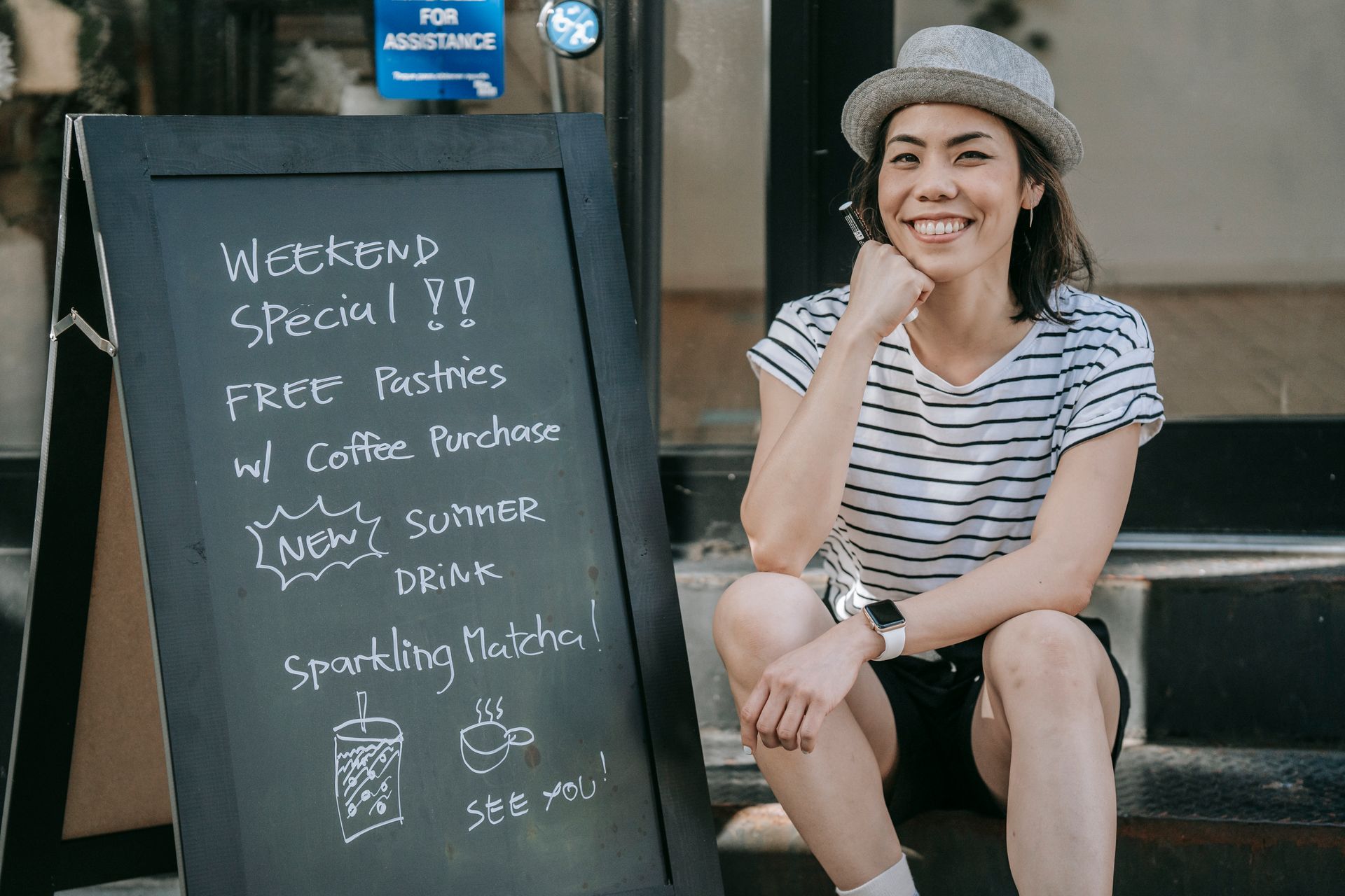 Asian woman enjoying a casual moment sitting beside chalkboard menu outside cafe.