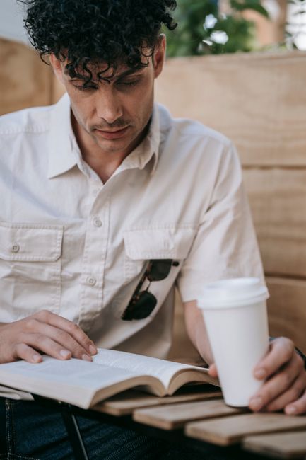 Man reading a book outdoors at a cafe while holding a coffee cup. Casual setting with natural ambiance. Man reading a book outdoors at a cafe while holding a coffee cup. Casual setting with natural ambiance.