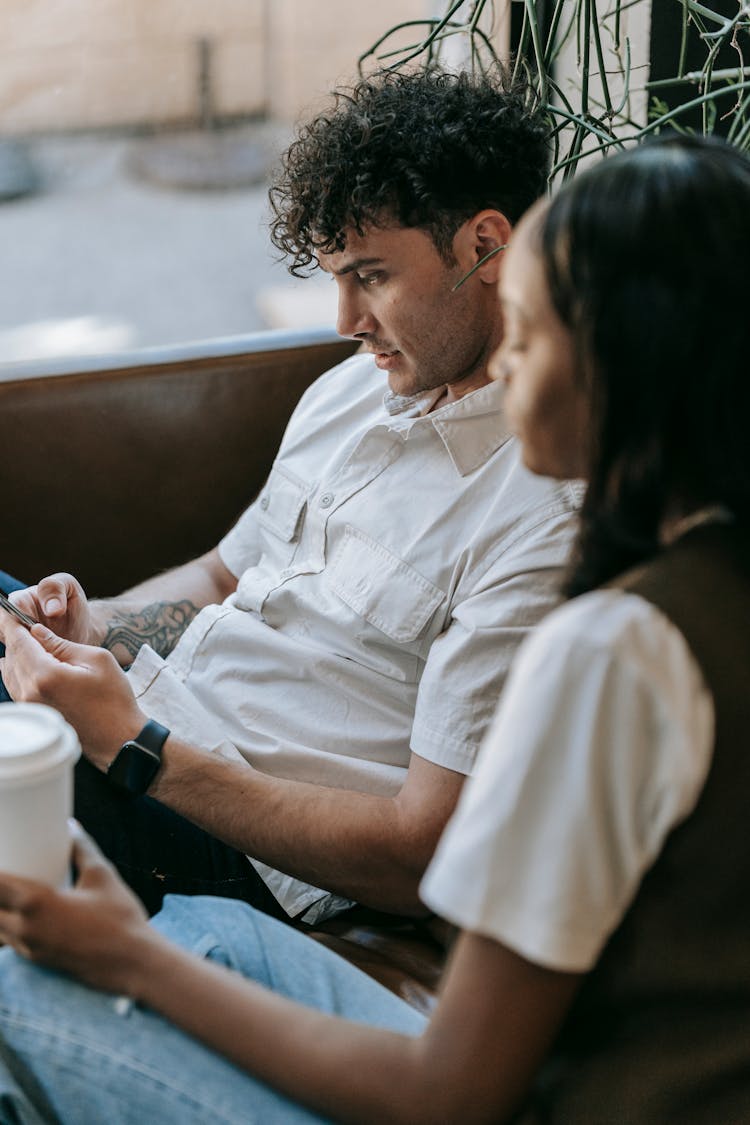 Woman Sitting And Holding A Disposable Coffee Cup And Man Sitting Next To Her Using His Phone 