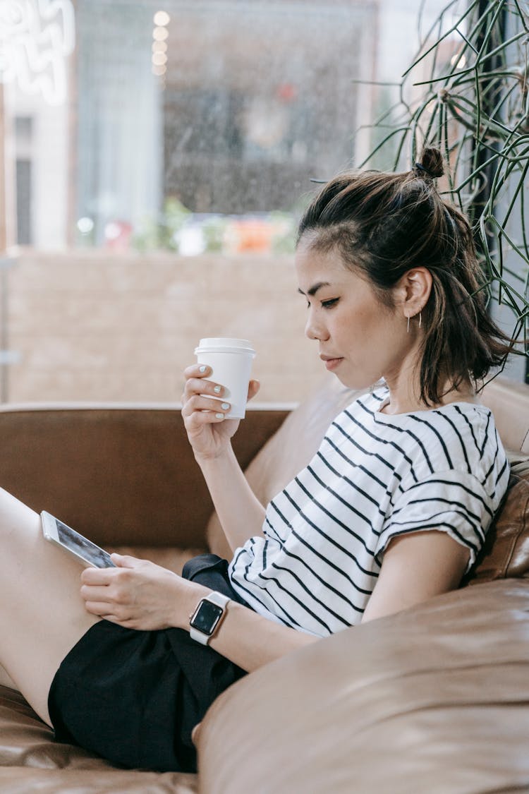 A Woman Using A Tablet While Holding A Coffee On A Couch