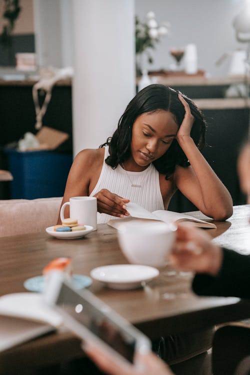 Woman in White Tank Top Sitting at the Table Reading a Book