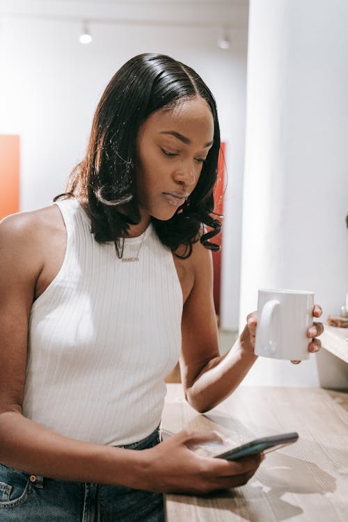 Woman in White Tank Top Holding White Mug While Using a Smart Phone