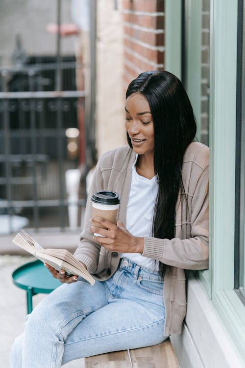 Woman in Brown Cardigan and Blue Denim Jeans Sitting on Bench Holding a Coffee Cup and Book