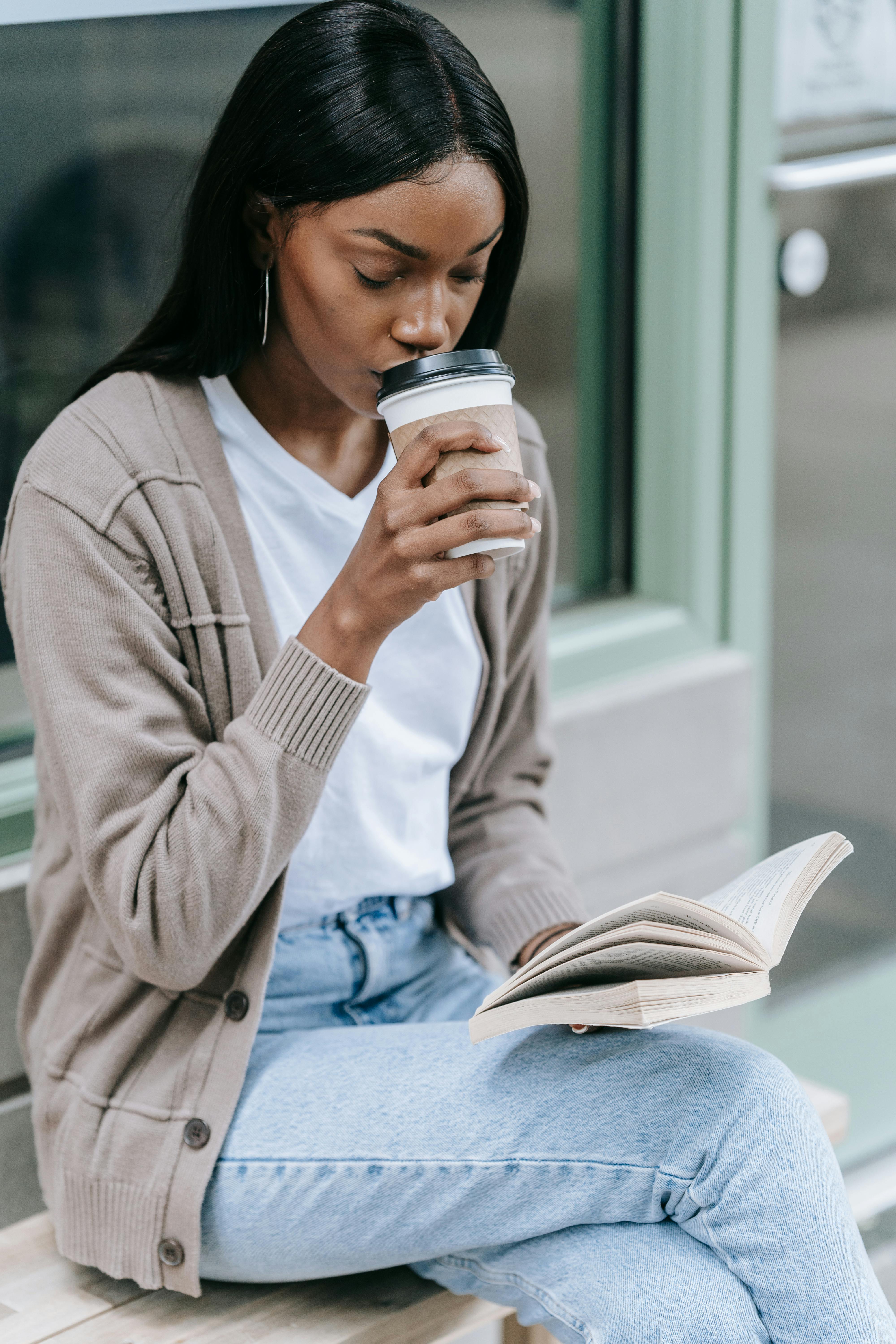 Woman sipping coffee while reading a book outdoors. Casual and relaxed scene.