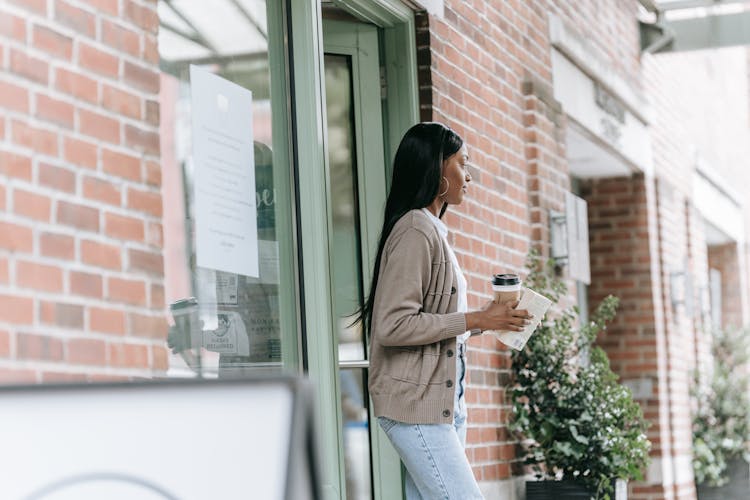 A Woman Walking Out Of A Cafe Holding A Book And A Cup Of Coffee