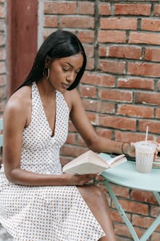 Woman reading a book with iced drink, sitting by a brick wall.