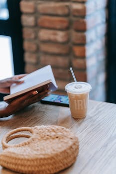 A person reading a book beside an iced coffee in a cozy cafe setting.