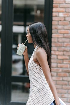 A woman in a polka dot top sips from a disposable cup while walking downtown.