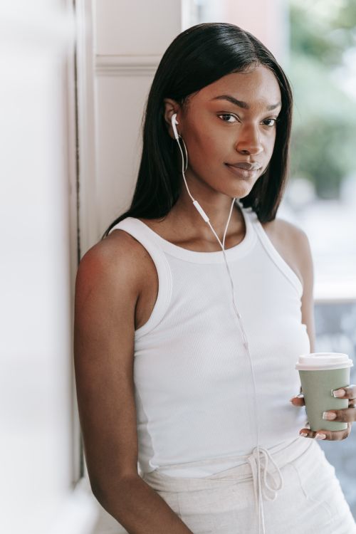 Free Confident woman in white tank top holding a coffee cup and listening to music with earphones. Stock Photo