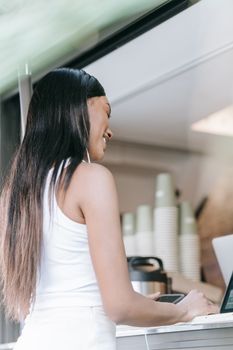 A woman with long black hair uses a touchscreen at a coffee stand on a sunny day.