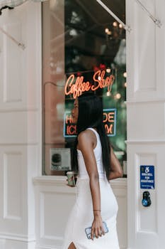 Black woman in white dress holding coffee and smartphone outside coffee shop.