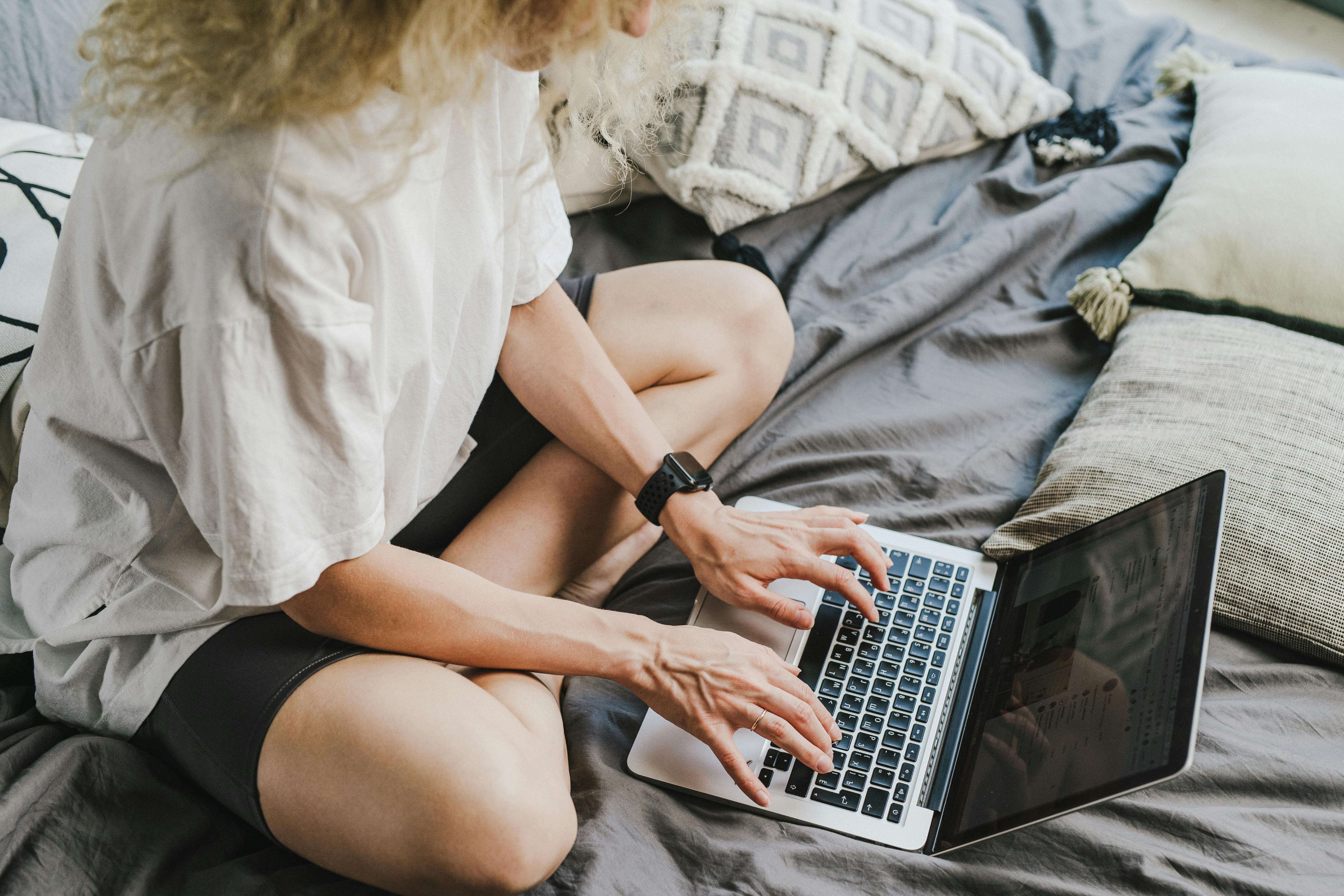 Woman typing on a laptop while sitting comfortably on a bed with pillows.