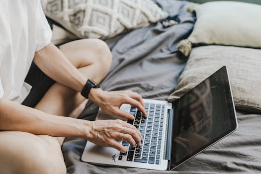 Person working on a laptop while sitting on a bed, showcasing a home office lifestyle.