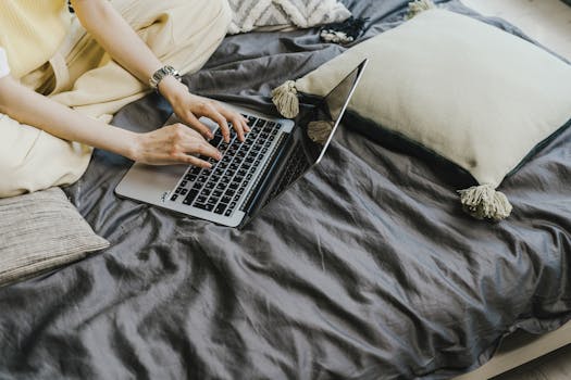 Person typing on a laptop while sitting on a bed with pillows. Perfect for remote work themes.