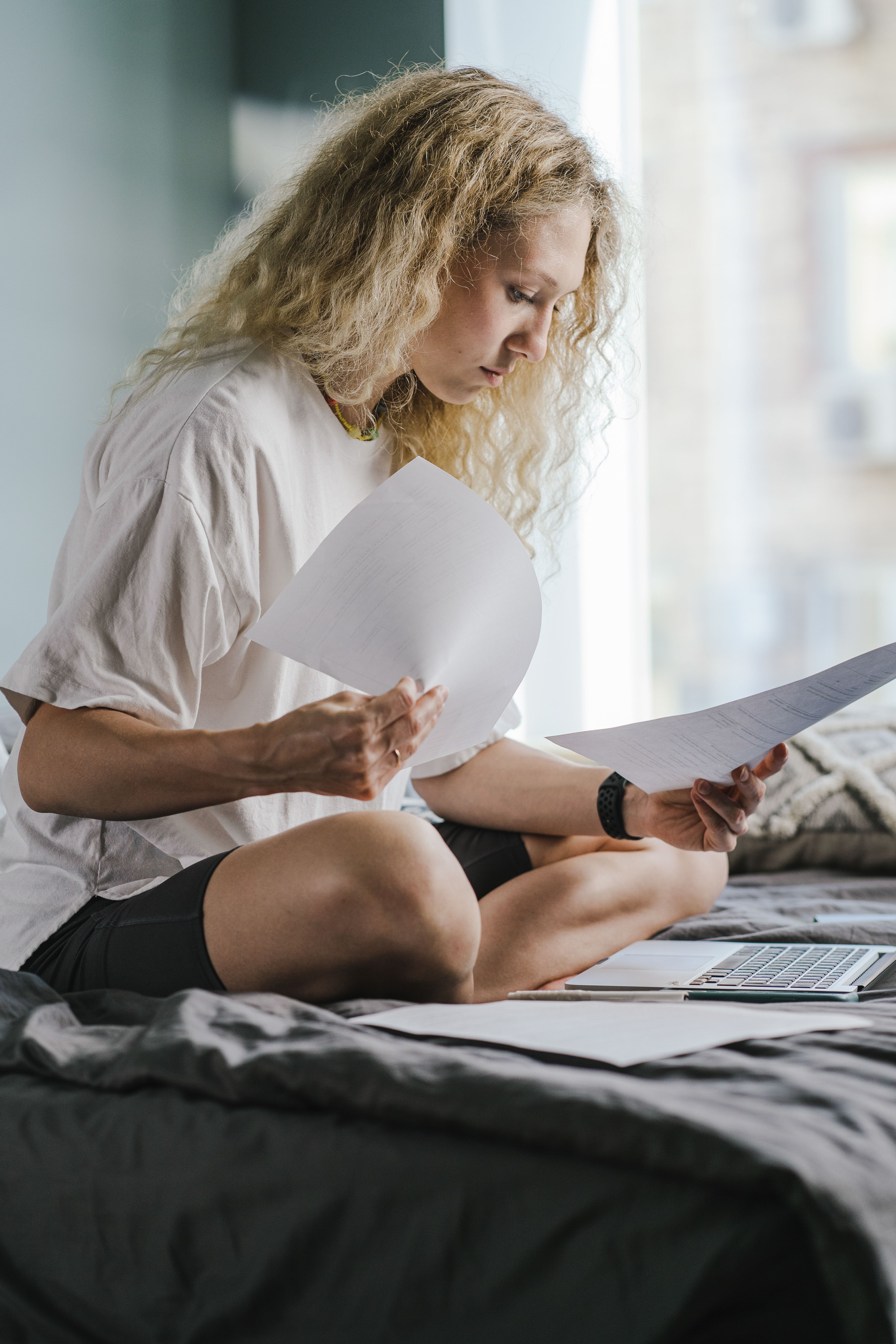 Focused woman reviewing documents while working indoors, sitting cross-legged on the bed.