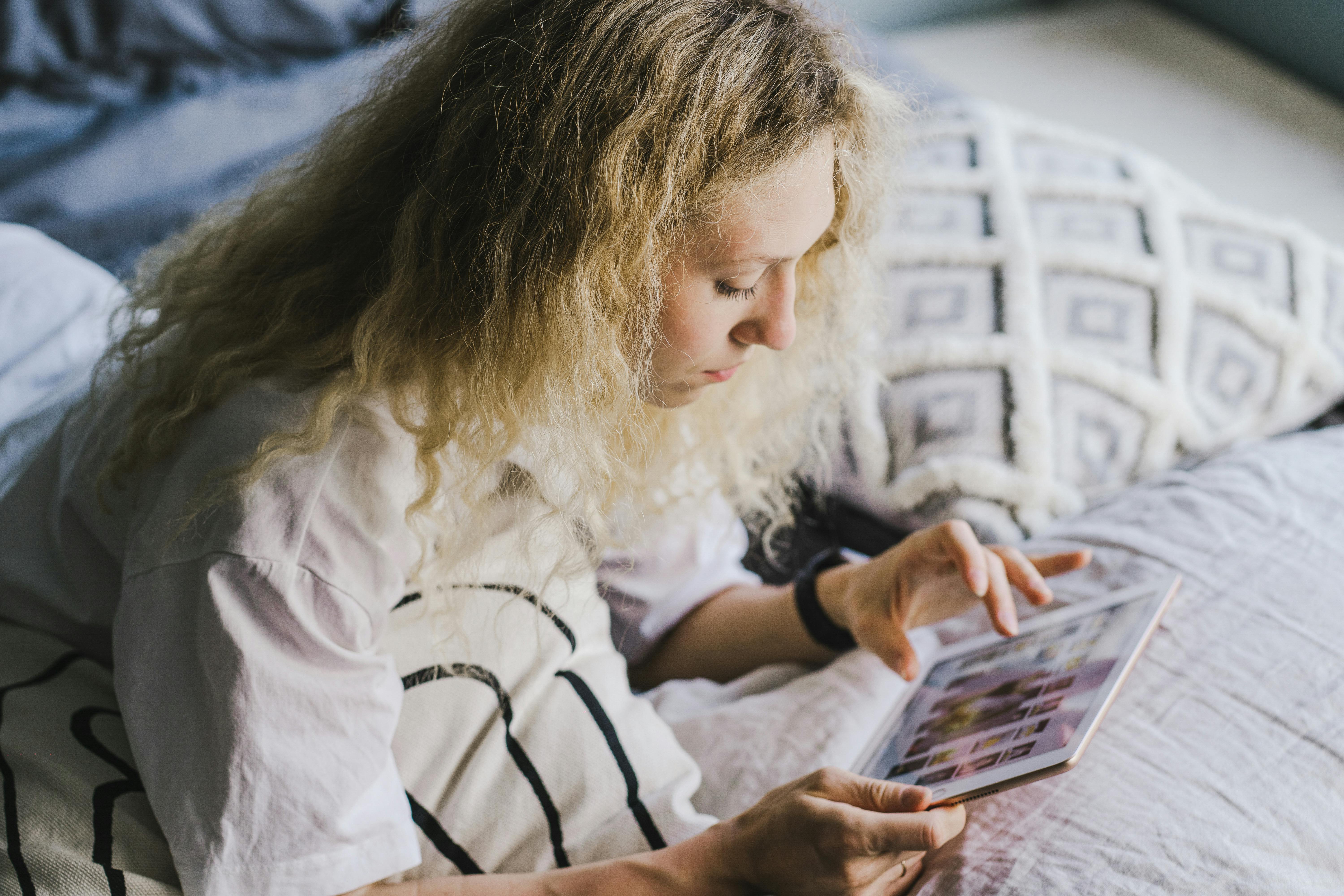 A young woman sits on her bed scrolling on a tablet, showcasing modern relaxation and technology.