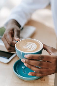 Close-up of hands holding a latte in a blue cup with a leaf pattern, placed indoors.
