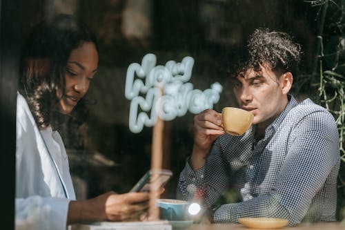 Man in Black and White Checkered Button Up Shirt Drinking from Brown Ceramic Mug Beside a Woman Using Cellphone