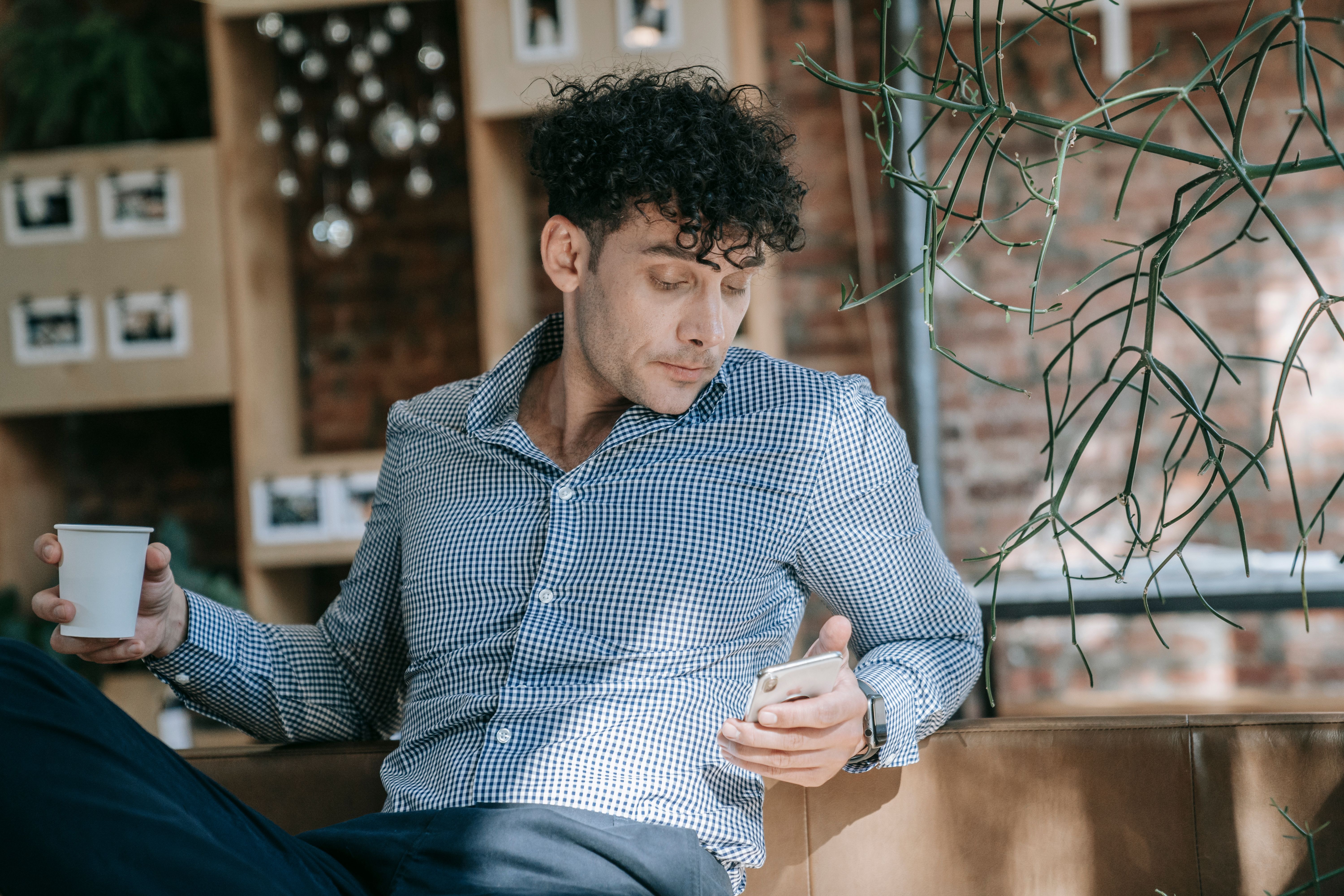 Man in stylish attire enjoying coffee while checking phone in a trendy cafe setting.