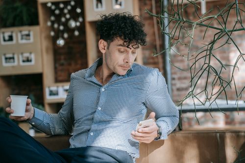 Free Man in stylish attire enjoying coffee while checking phone in a trendy cafe setting. Stock Photo