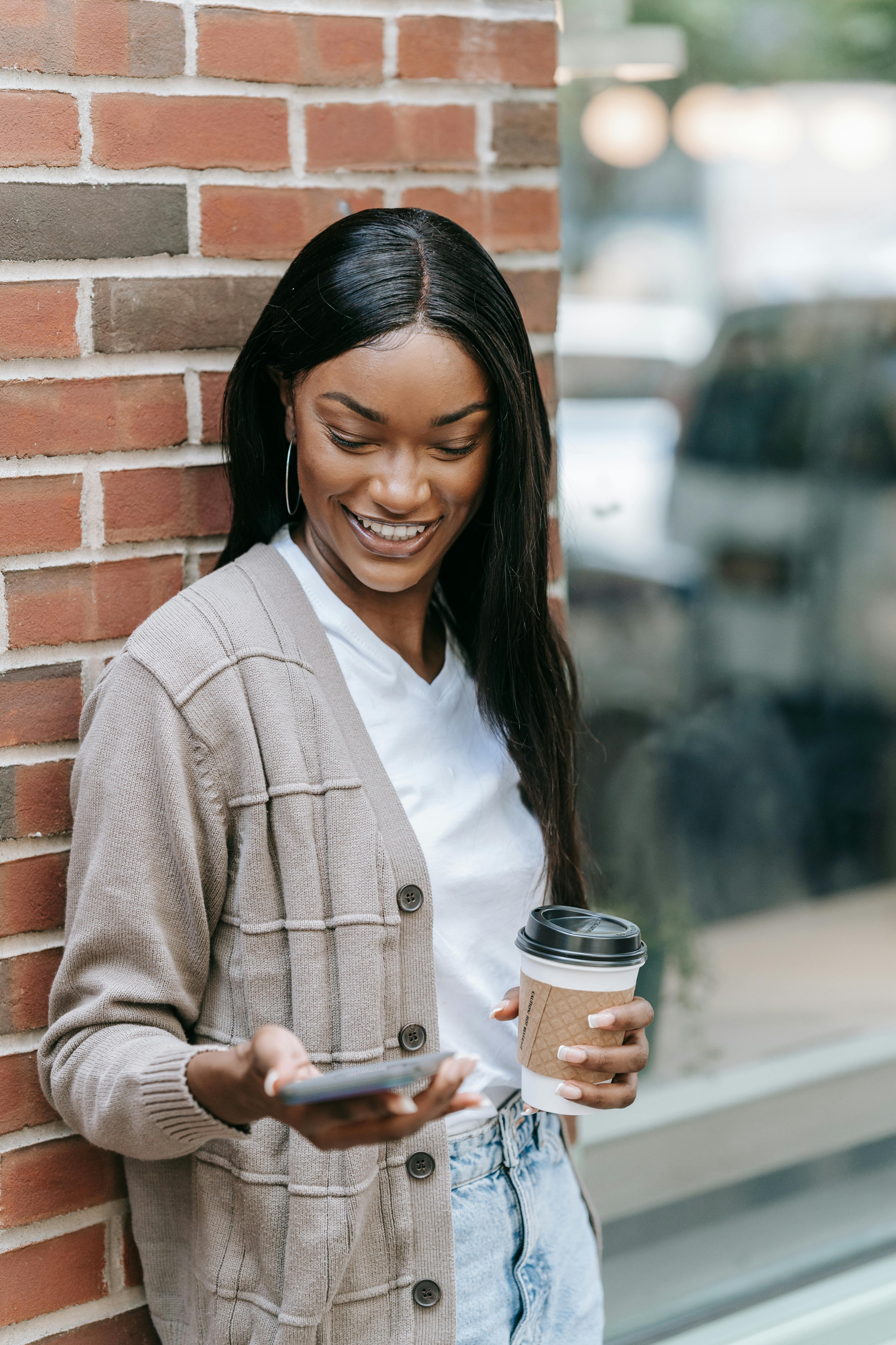 Smiling woman stands by a brick wall, sipping coffee and checking phone outdoors.