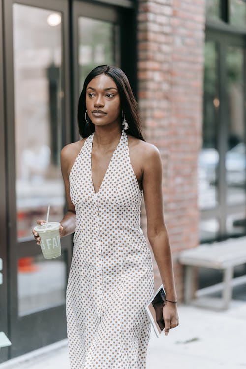 Woman in White and Brown Polka Dot Halter Dress Walking on Sidewalk Holding a Cup and Digital Tablet