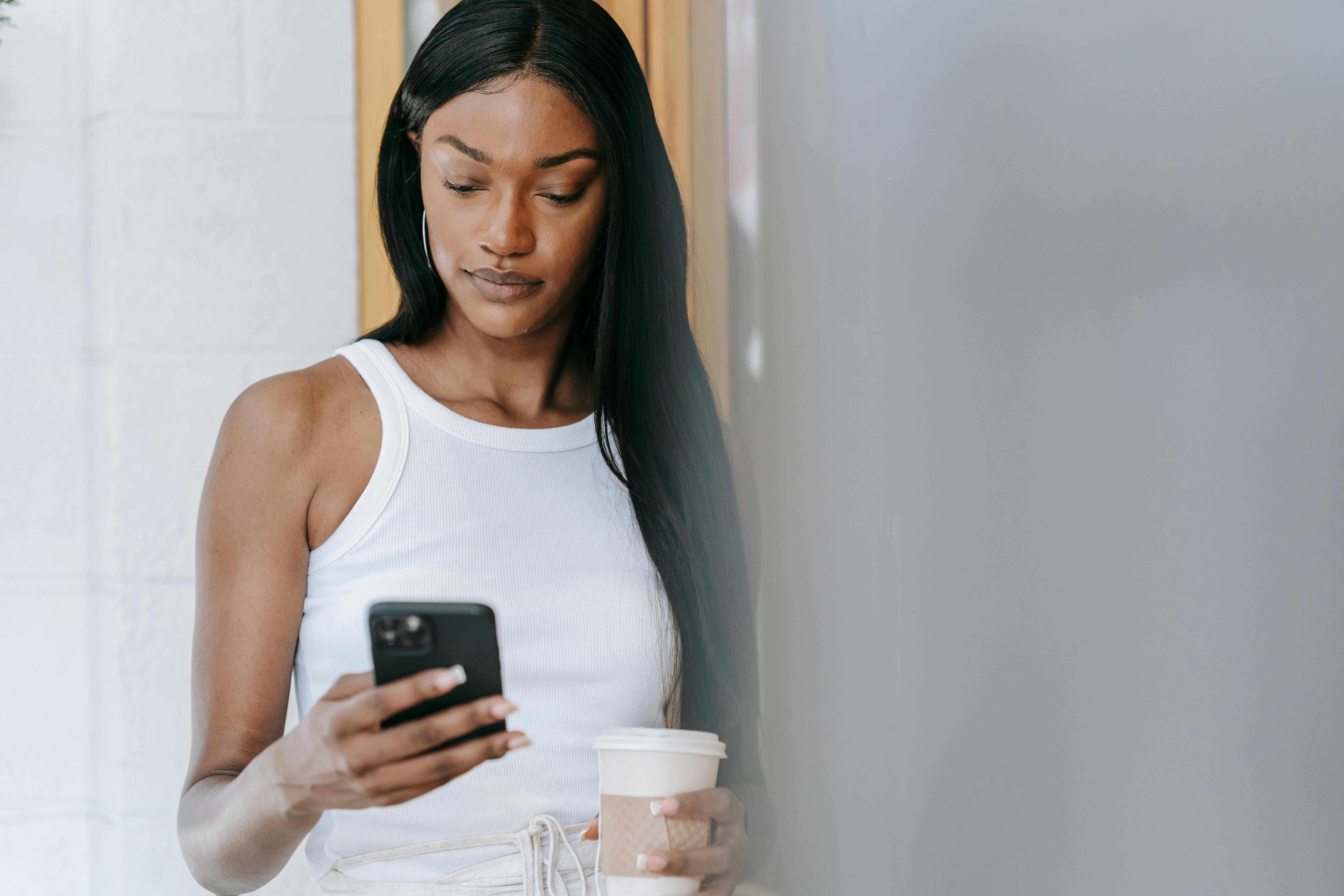Young woman using her smartphone while holding a coffee cup indoors.